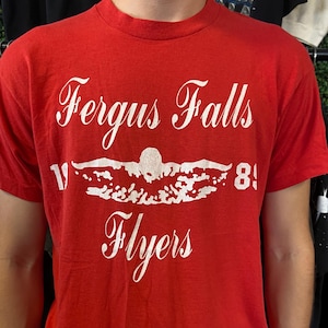 Puede incluir: Camiseta roja con texto blanco que dice "Fergus Falls Flyers" encima de un gr&aacute;fico de nadador y el n&uacute;mero "81". La camiseta tiene mangas cortas y cuello redondo.