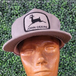 May include: A gray corduroy baseball cap with a flat brim. The cap features a rectangular patch with the John Deere logo, a black deer silhouette, and the brand name in white lettering.