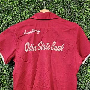 Może przedstawiać: Bordowa koszula z krótkim rękawem z białym wykończeniem na rękawach i kołnierzu. Tył koszuli zdobi haftowany napis "Sandberg" nad "Odin State Bank" w stylu vintage.