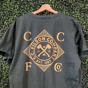 May include: Dark green t-shirt with a gold diamond-shaped graphic on the back. The graphic features crossed axes and the text "C.C. FILSON COMPANY EST. 1897". The shirt is hanging on a wooden hanger.