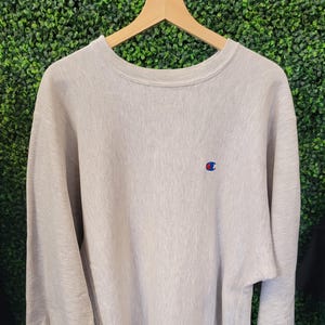 Puede incluir: Sudadera Champion gris claro con un pequeño logotipo en el pecho y la manga. La sudadera de cuello redondo cuelga de una percha de madera sobre un fondo verde. La sudadera tiene puños y dobladillo acanalados.