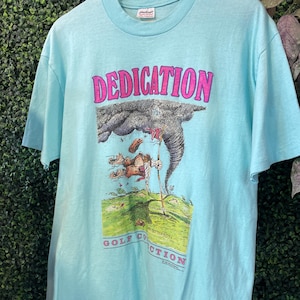 May include: Light blue t-shirt with the word "DEDICATION" in pink block letters above a golf-themed graphic. The graphic depicts a golfer battling a tornado. The words "GOLF COLLECTION" are printed below the graphic.