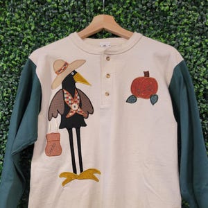 Pode incluir: Camiseta de manga comprida creme com uma aplicação de corvo e abóbora. O corvo usa um chapéu e uma cruz decorativa. As mangas são verde escuro. A camisa tem decote abotoado e uma aplicação de abóbora.