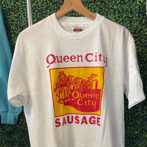 May include: White t-shirt with a graphic featuring the words "Queen City Sausage" in red and yellow. The design includes an illustration of a person and buildings. The shirt is hanging on a wooden hanger.