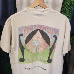 May include: Cream-coloured t-shirt with a colourful graphic of a mountain scene. The text reads "Yonder Mountain String Band" and "Nederland, Colorado." The design includes musical notes and a figure playing an instrument.