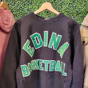 以下が含まれることがあります： 胸元に白い縁取りの大きな緑色の文字で「EDINA BASKETBALL」と書かれた、黒い長袖のクルーネックトレーナー。左袖口付近には、小さなChampionのロゴ刺繍が施されています。