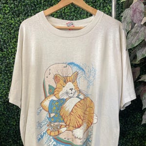 May include: Cream-colored t-shirt featuring a cartoon cat on a surfboard with blue waves. The cat is orange and white, wearing a blue and gold outfit. Text on the shirt reads "U.B. Koo Minnesota."