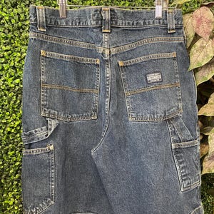 May include: Dark blue denim shorts with multiple pockets and a "STRUCTURE" label. The shorts have a classic design with visible stitching and a button and zipper closure. The shorts are hanging on a wooden hanger.