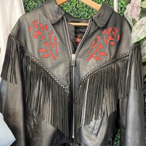 May include: Black leather jacket with red rose embroidery on the shoulders. The jacket has long fringe along the chest and sleeves, a zip closure, and side lacing details. The jacket is hanging on a wooden hanger.