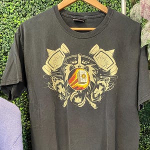 May include: Black t-shirt with a gold and red graphic design featuring hammers, a sun, and other symbols. The shirt is hanging on a wooden hanger against a green background.