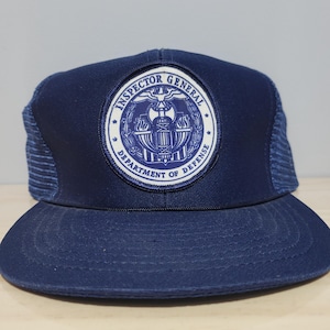May include: A navy blue trucker cap with a curved peak and mesh back. The front displays a circular patch with the text "INSPECTOR GENERAL DEPARTMENT OF DEFENSE" in white and blue.