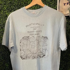 May include: Light blue t-shirt with the text "PARTICIPANT CALIFORNIA'S 1987 TRINITY COUNTY FIRE GAMES" and a map graphic. The shirt also features a "1st PLACE" emblem. The shirt is hanging on a wooden hanger.