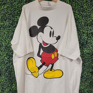 May include: White t-shirt featuring a large graphic of Mickey Mouse. Mickey is depicted in his classic pose, wearing red shorts, yellow shoes, and black gloves. The shirt is hanging on a wooden hanger against a green textured background.