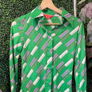 May include: A long-sleeve button-down shirt in a vibrant green hue featuring a geometric pattern of white and multi-colored striped rectangles. The shirt has a pointed collar, a front button placket, and a red label at the neck reading Trousers.