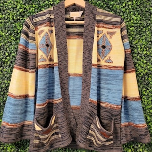 May include: A long-sleeve open-front cardigan sweater featuring a horizontal striped pattern in shades of charcoal gray, light blue, and cream. The garment includes two front patch pockets and a diamond-shaped geometric motif on the chest area.