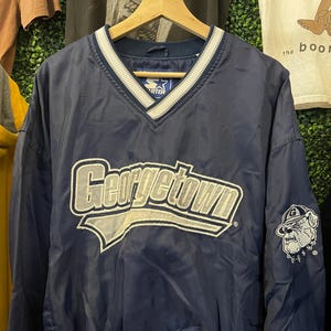 May include: A navy blue Starter brand pullover windbreaker jacket featuring a V-neck collar with white and navy stripes. The front displays the word Georgetown in white and grey embroidery, while the left sleeve features a bulldog mascot logo.