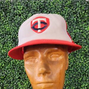 May include: A white and red baseball cap with a red brim and a logo featuring a stylized "TC" in red and navy blue. The cap is displayed on a wooden mannequin head against a backdrop of green foliage.