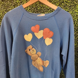 May include: A blue long-sleeve sweatshirt featuring a graphic print of a brown teddy bear holding a cluster of four heart-shaped balloons in shades of red, orange, and yellow. The garment has a ribbed crew neckline, cuffs, and a ribbed hem.