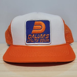 Könnte beinhalten: Orange-weiße Trucker-Cap mit einem quadratischen Aufnäher mit der Aufschrift "DALUM'S UTILITY EQUIP." Der Aufnäher hat einen blauen Hintergrund und ein orangefarbenes "D"-Logo. Die Cap hat eine weiße Front, einen orangefarbenen Schirm und ein orangefarbenes Mesh-Rückenteil.