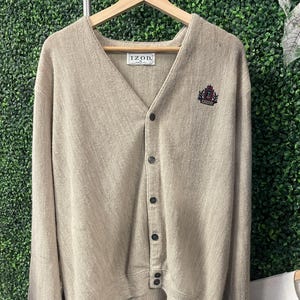 May include: A beige IZOD cardigan sweater with a V-neck, long sleeves, and button-up front. The sweater has a small emblem on the upper left chest and is hanging on a wooden hanger.