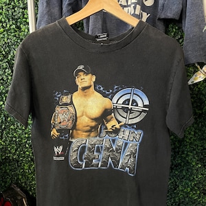 Puede incluir: Camiseta negra con un gráfico del luchador John Cena sosteniendo un cinturón de campeonato. El diseño incluye el texto "JOHN CENA" en fuente azul y blanca, y un gráfico de diana. La camiseta está colgada en una percha de madera.
