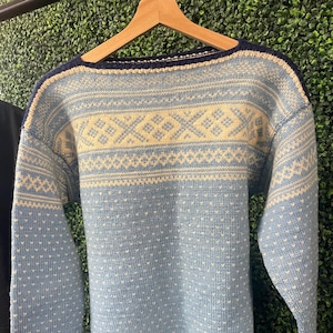 May include: A light blue sweater with a cream-colored geometric pattern across the chest and shoulders. The sweater has long sleeves and a crew neck with navy blue trim. Small white dots are evenly spaced across the light blue knit.