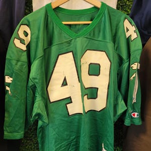 Peut inclure: Maillot de football vert avec le numéro 49 en blanc sur le devant et les manches. Le maillot a un col en V et est suspendu à un cintre en bois. Le maillot porte le logo Champion sur la manche.