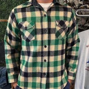 May include: A green, black, and cream plaid button-up shirt with two chest pockets. The shirt has a black collar and black buttons. The long-sleeved shirt is made of a thick, textured fabric.