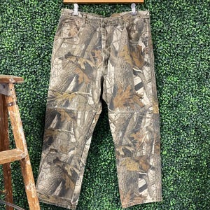 May include: Camouflage trousers hanging on a wooden hanger. The trousers feature a brown, green, and tan woodland camouflage pattern. The trousers have multiple pockets and a button and zip closure. The trousers are made of a durable fabric.