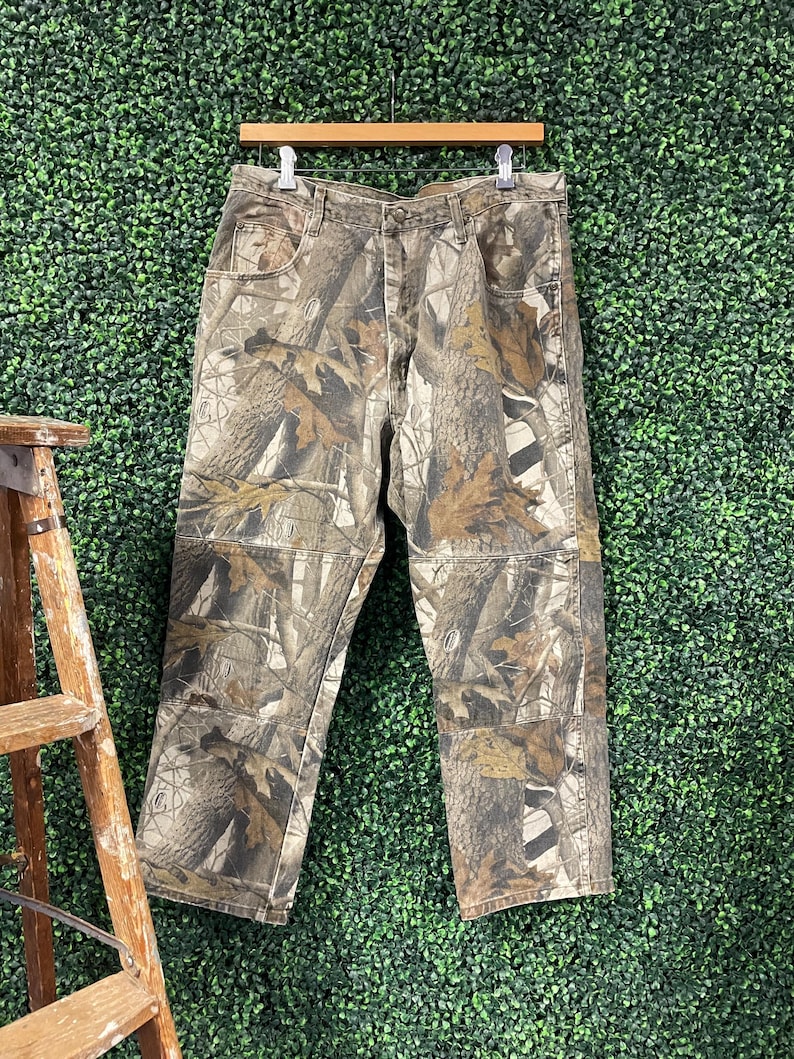May include: Camouflage trousers hanging on a wooden hanger. The trousers feature a brown, green, and tan woodland camouflage pattern. The trousers have multiple pockets and a button and zip closure. The trousers are made of a durable fabric.