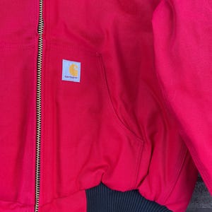 Pode incluir: Casaco Carhartt vermelho com bainha e punhos canelados pretos. O casaco tem fecho frontal, um bolso e o emblema com o log&oacute;tipo Carhartt. Uma etiqueta amarela est&aacute; presa ao fecho.
