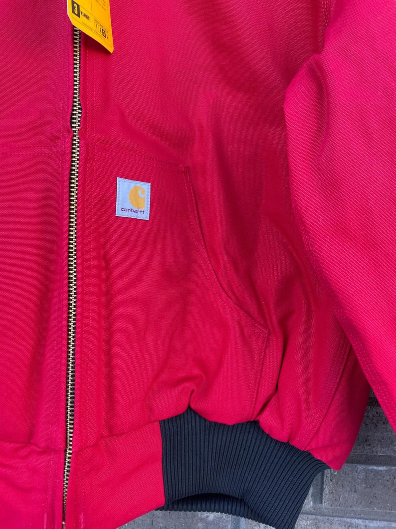 Pode incluir: Casaco Carhartt vermelho com bainha e punhos canelados pretos. O casaco tem fecho frontal, um bolso e o emblema com o log&oacute;tipo Carhartt. Uma etiqueta amarela est&aacute; presa ao fecho.