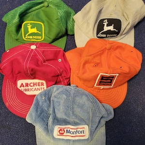Op de afbeelding: Een verzameling van zes baseballpetten in verschillende kleuren, waaronder groen, rood, oranje en blauw. Elke pet heeft een ander logo of merknaam, zoals John Deere, Archer Lubricants, Schneider en Monfort. De petten zijn gemaakt van verschillende materialen.