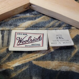 May include: Close-up of a camouflage-patterned shirt with the Woolrich label. The label reads "Since 1830, Woolrich, Original Outdoorwear." The shirt is labeled "Mens 2XL" and made of 100% cotton in Mexico. The shirt is hanging on a wooden hanger.
