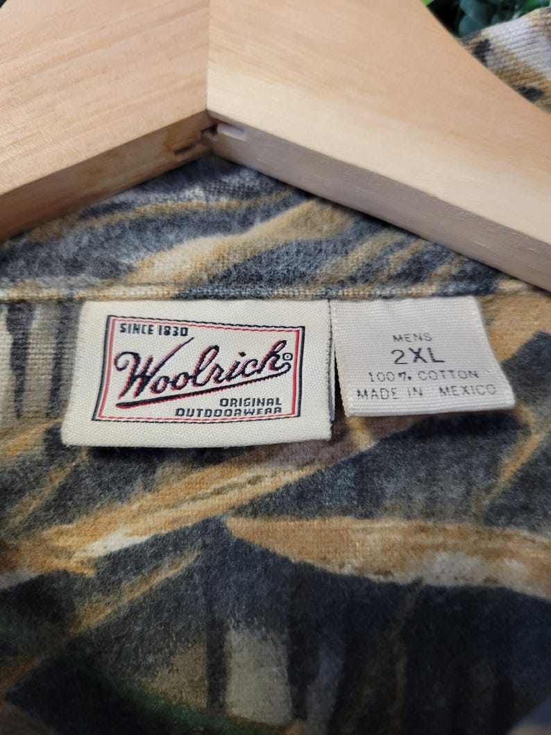 May include: Close-up of a camouflage-patterned shirt with the Woolrich label. The label reads "Since 1830, Woolrich, Original Outdoorwear." The shirt is labeled "Mens 2XL" and made of 100% cotton in Mexico. The shirt is hanging on a wooden hanger.