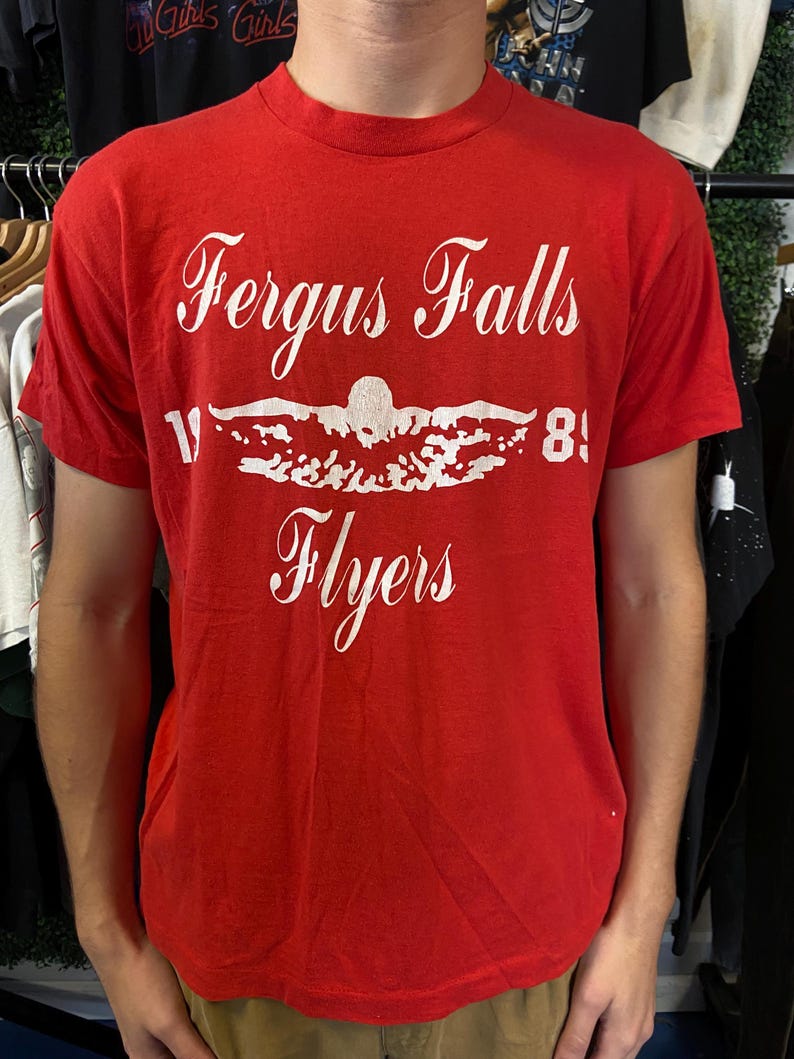 Puede incluir: Camiseta roja con texto blanco que dice "Fergus Falls Flyers" encima de un gr&aacute;fico de nadador y el n&uacute;mero "81". La camiseta tiene mangas cortas y cuello redondo.