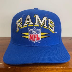 Puede incluir: Una gorra de béisbol azul real con la palabra RAMS bordada en blanco con contornos amarillos y negros. Debajo del texto hay un logotipo de escudo de la NFL bordado en rojo, blanco y azul, flanqueado por dos rayos amarillos horizontales.