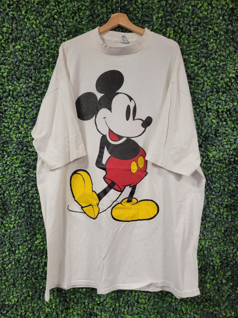 May include: White t-shirt featuring a large graphic of Mickey Mouse. Mickey is depicted in his classic pose, wearing red shorts, yellow shoes, and black gloves. The shirt is hanging on a wooden hanger against a green textured background.