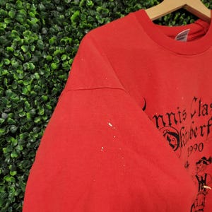 May include: Red t-shirt with black text and a graphic of a person in traditional attire. The text reads "Tennis Classic Oktoberfest 1990". The shirt is hanging on a wooden hanger against a green background.