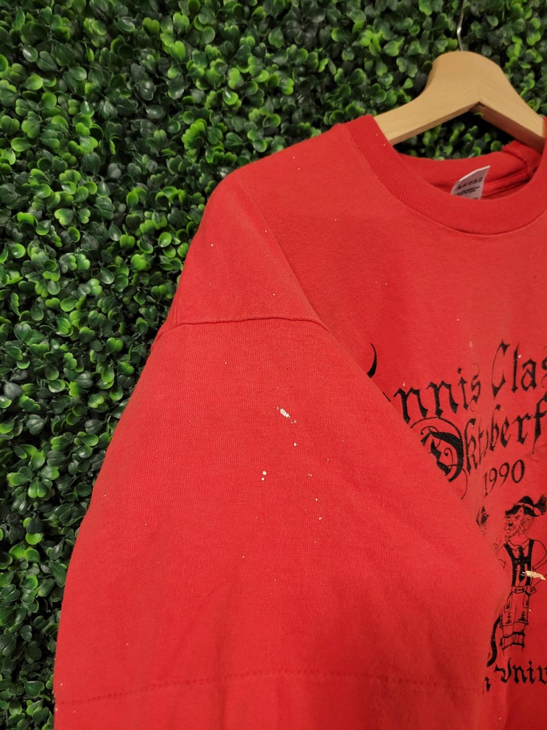 May include: Red t-shirt with black text and a graphic of a person in traditional attire. The text reads "Tennis Classic Oktoberfest 1990". The shirt is hanging on a wooden hanger against a green background.