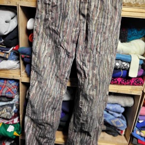 May include: Camouflage-patterned pants in shades of brown, green, and gray. The pants have reinforced knee patches and a classic workwear design. The fabric has a vertical, tree-like pattern, ideal for outdoor activities or a unique fashion statement.