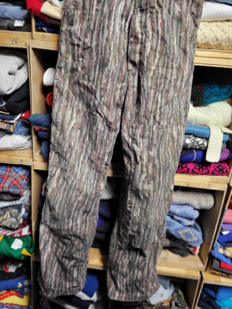 May include: Camouflage-patterned pants in shades of brown, green, and gray. The pants have reinforced knee patches and a classic workwear design. The fabric has a vertical, tree-like pattern, ideal for outdoor activities or a unique fashion statement.