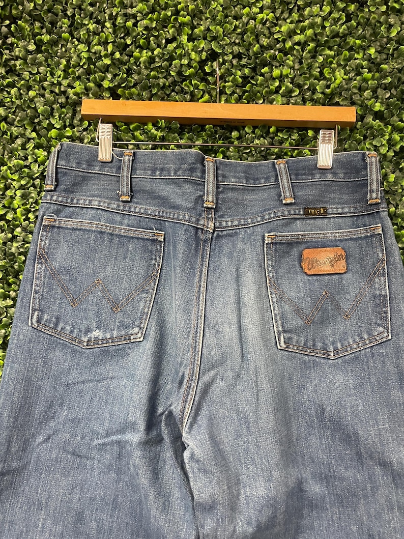 May include: Close-up of blue denim jeans with the Wrangler logo on the back pocket. The jeans are hanging on a wooden hanger against a green background. The jeans have two back pockets and a classic design.