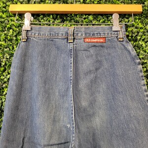 May include: Close-up of blue denim jeans hanging on a wooden hanger. The jeans have a straight leg cut and a red tag that reads "R.D. SIMPSON". The background is a wall of green foliage.