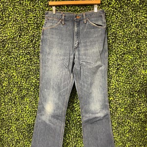 May include: Blue denim jeans with a classic five-pocket design, hanging on a wooden hanger. The jeans have a faded wash and a slight flare at the bottom, suggesting a vintage style. The background is a textured green.