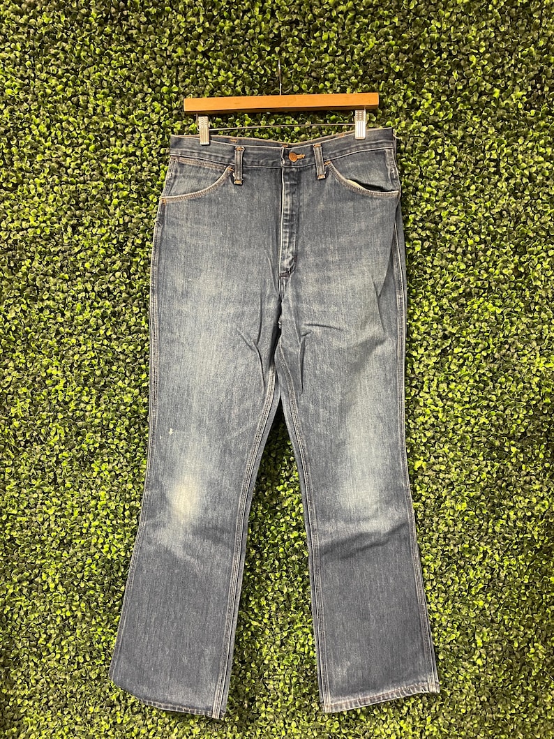 May include: Blue denim jeans with a classic five-pocket design, hanging on a wooden hanger. The jeans have a faded wash and a slight flare at the bottom, suggesting a vintage style. The background is a textured green.