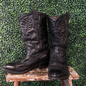 May include: A pair of black leather cowboy boots with decorative stitching. The boots are worn and sit on a small wooden stool against a green backdrop. The boots have a pointed toe and a tall shaft.