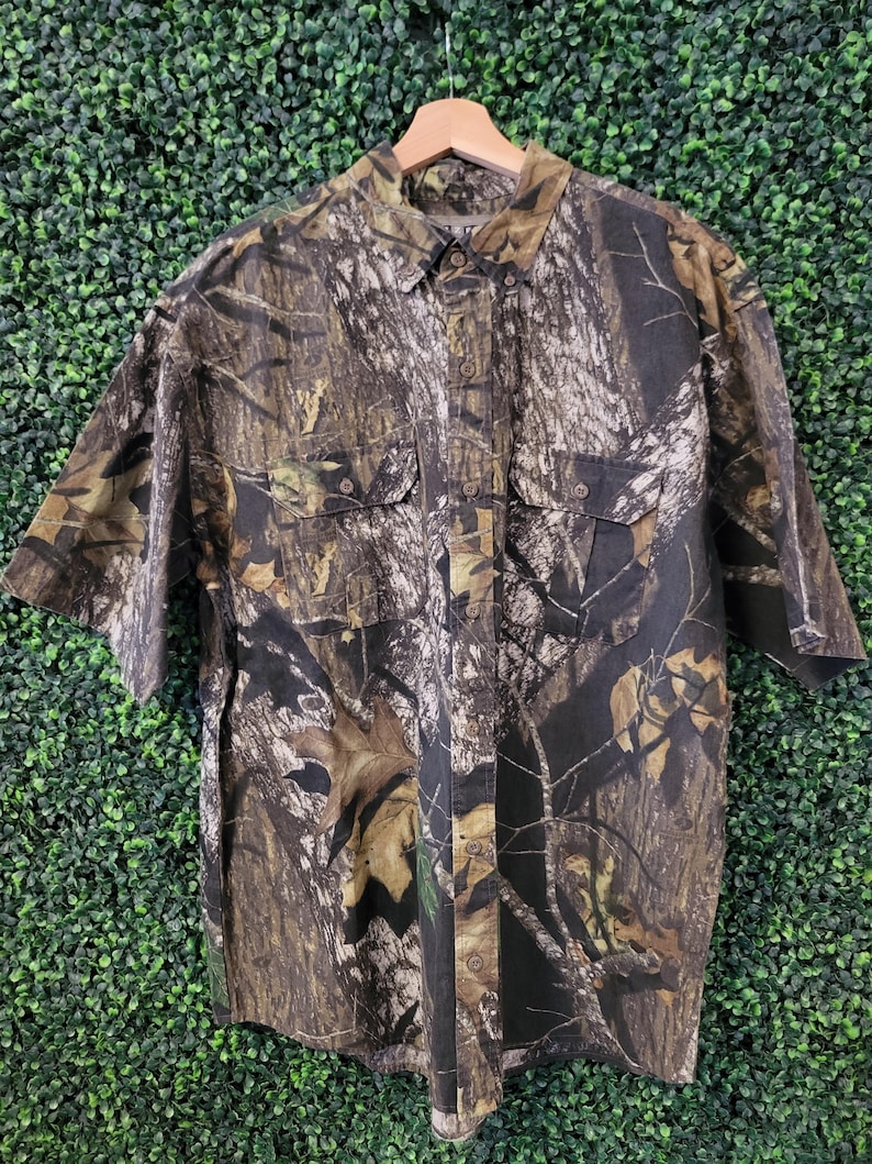 Op de afbeelding: Korte mouwen button-down shirt met een camouflagepatroon in bruin, groen en zwart. Het shirt heeft twee borstzakken en hangt aan een houten hanger. De stof lijkt licht van gewicht.