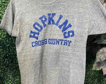 May include: A heather gray short-sleeve t-shirt with a crew neckline. The front features the blue text HOPKINS in an arched design above the blue text CROSS COUNTRY. The shirt is displayed on a wooden hanger against a green foliage background.