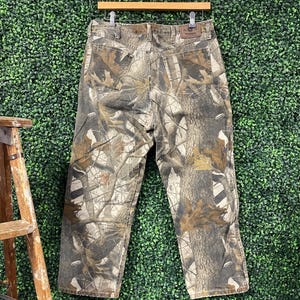 May include: Camouflage-patterned jeans hanging on a wooden hanger. The trousers feature a mix of brown, green, and tan colours, creating a realistic forest design. The jeans have pockets and a leather patch.